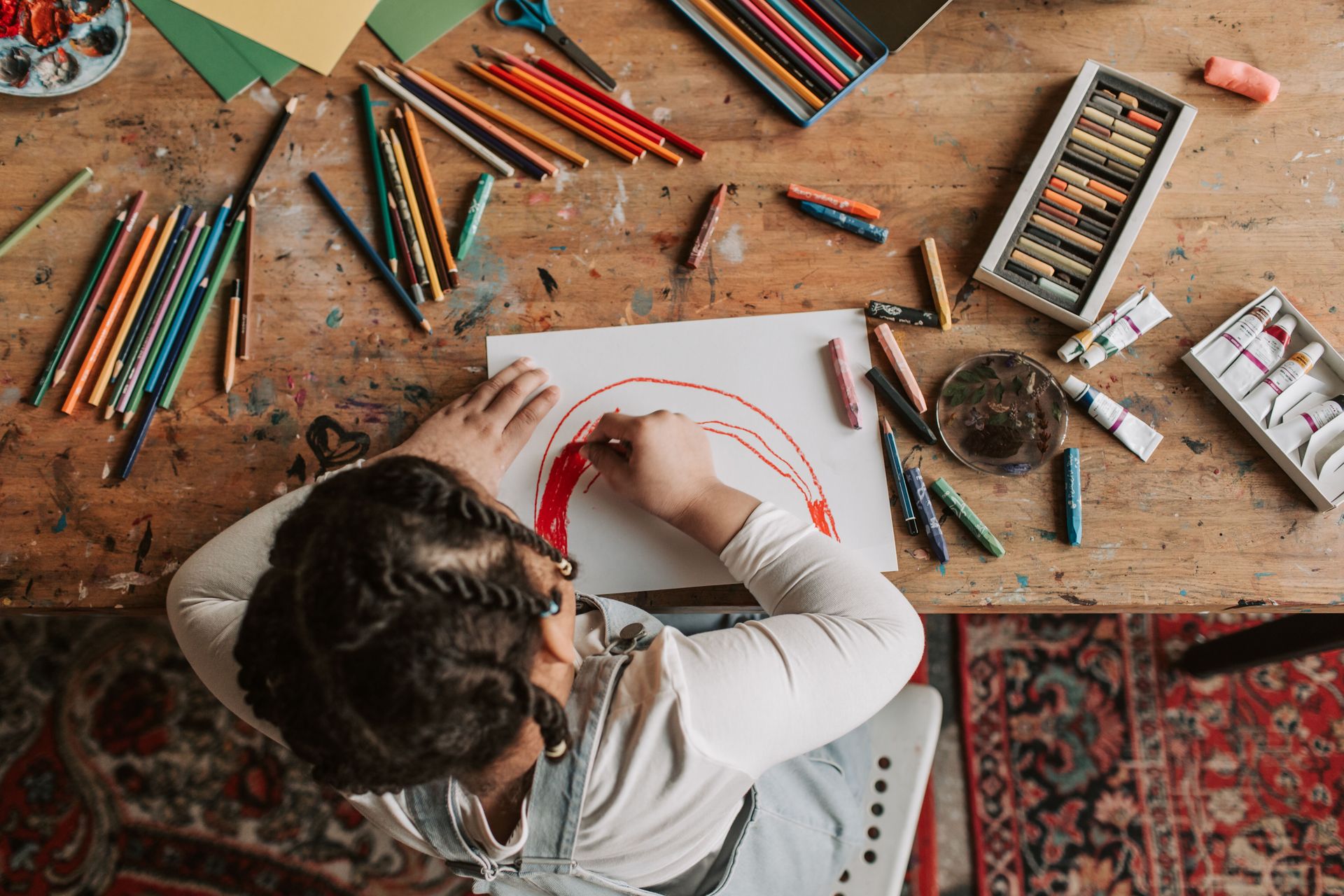 Child engaged in creative drawing with colored pencils, crayons, and paints at a wooden table.